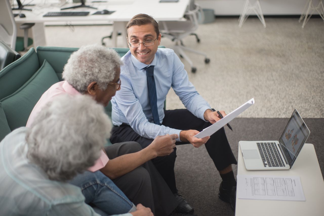 contact-img Consultant discussing financial plans with senior clients in a modern office setting, using documents and a laptop.